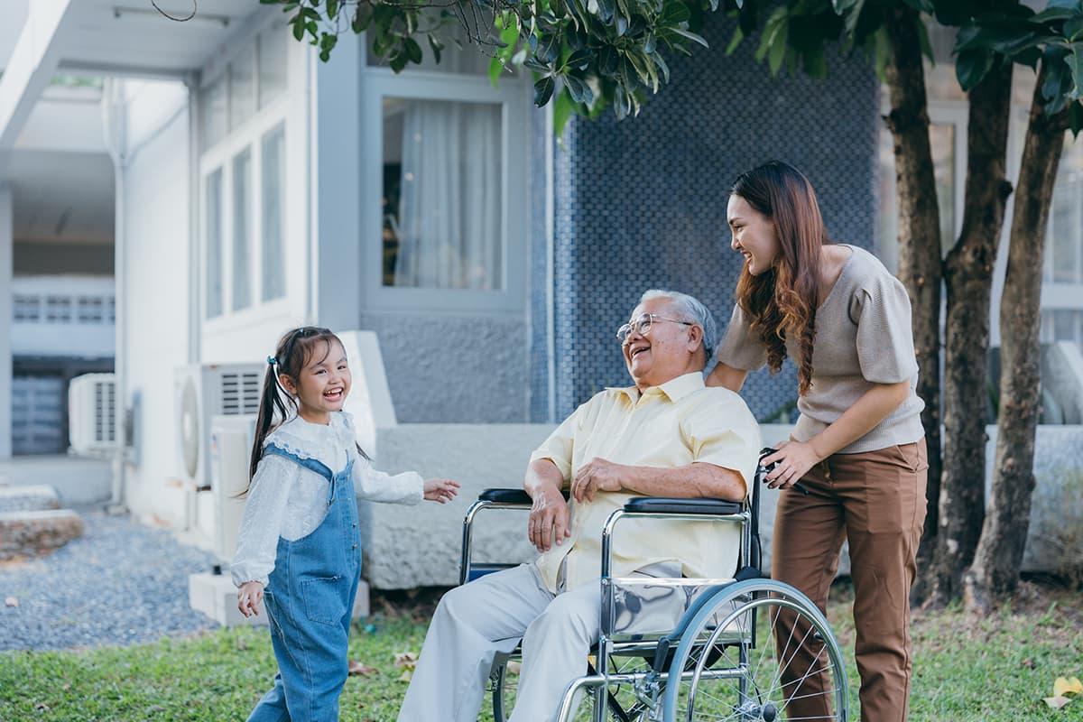 Social worker laughs along with a wheelchair bound elderly man and his grandaughter outside a house under a tree.